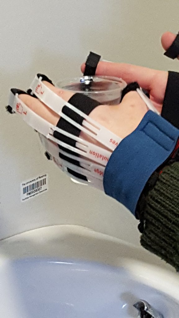 Two hands wearing gloves which simulate dexterity impairment holding a plastic cup full of water.