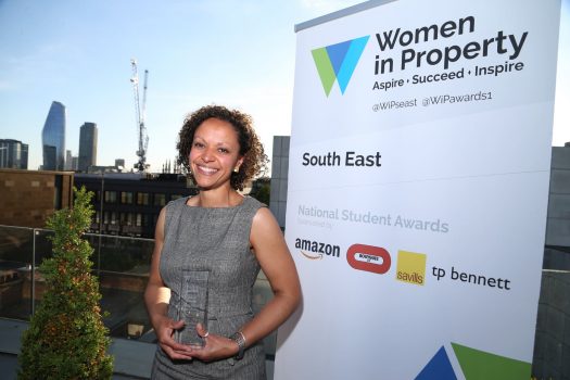 Image of Mary Utley, winner of the Women in Property South East Student Awards 2022. Mary is holding a glass award and stands behind a banner which has the name of the award on it.