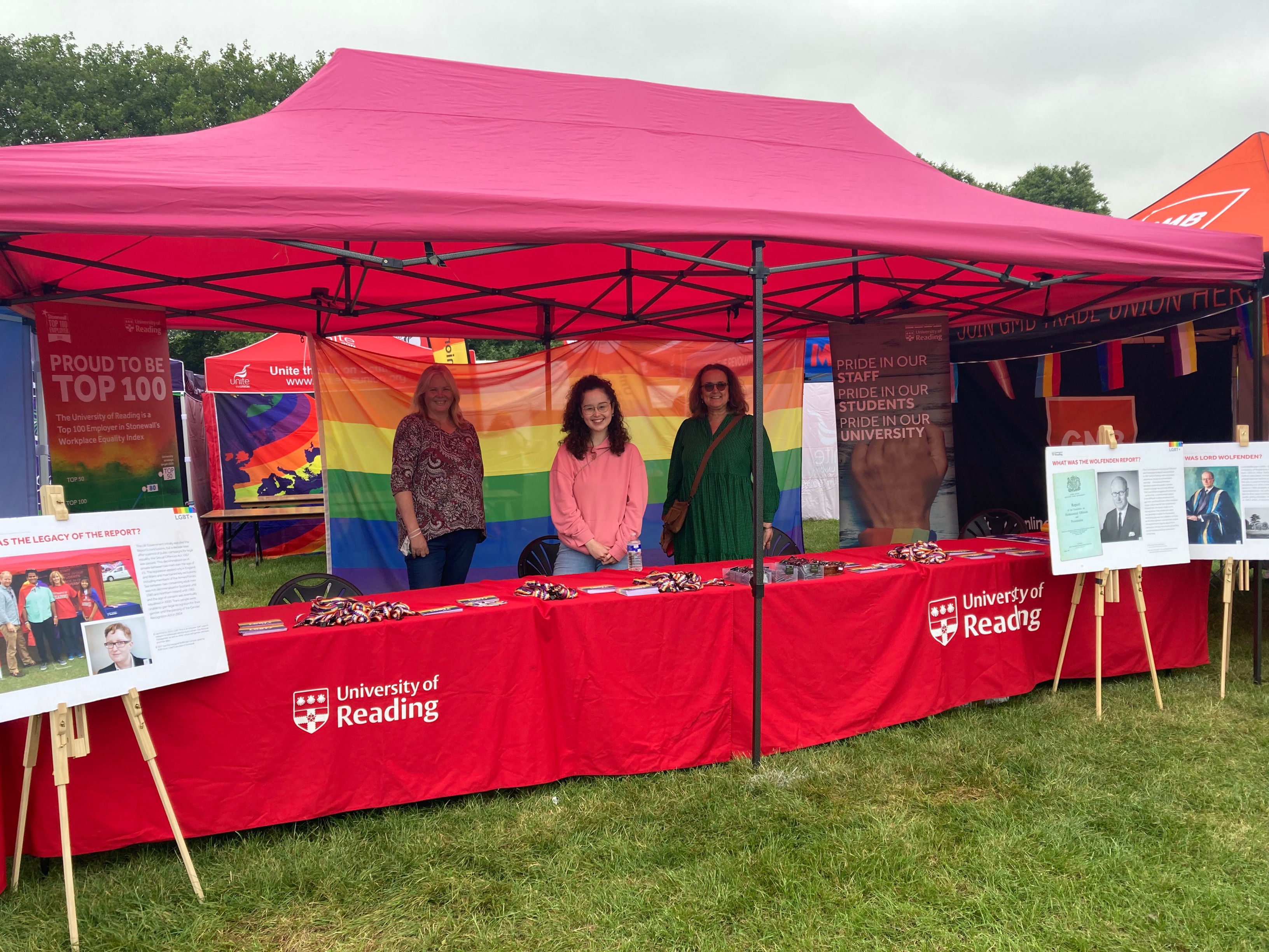  Professor Kat Bicknell, Head of the Department of Pharmacy, Nozomi Tolworthy, Diversity and Inclusion Advisor and Professor Carol Fuller stand under the gazebo at the University of Reading stall. They are standing in front of a table which has a red University of Reading table cloth and is covered with rainbow lanyards, postcards and pronoun badges. 