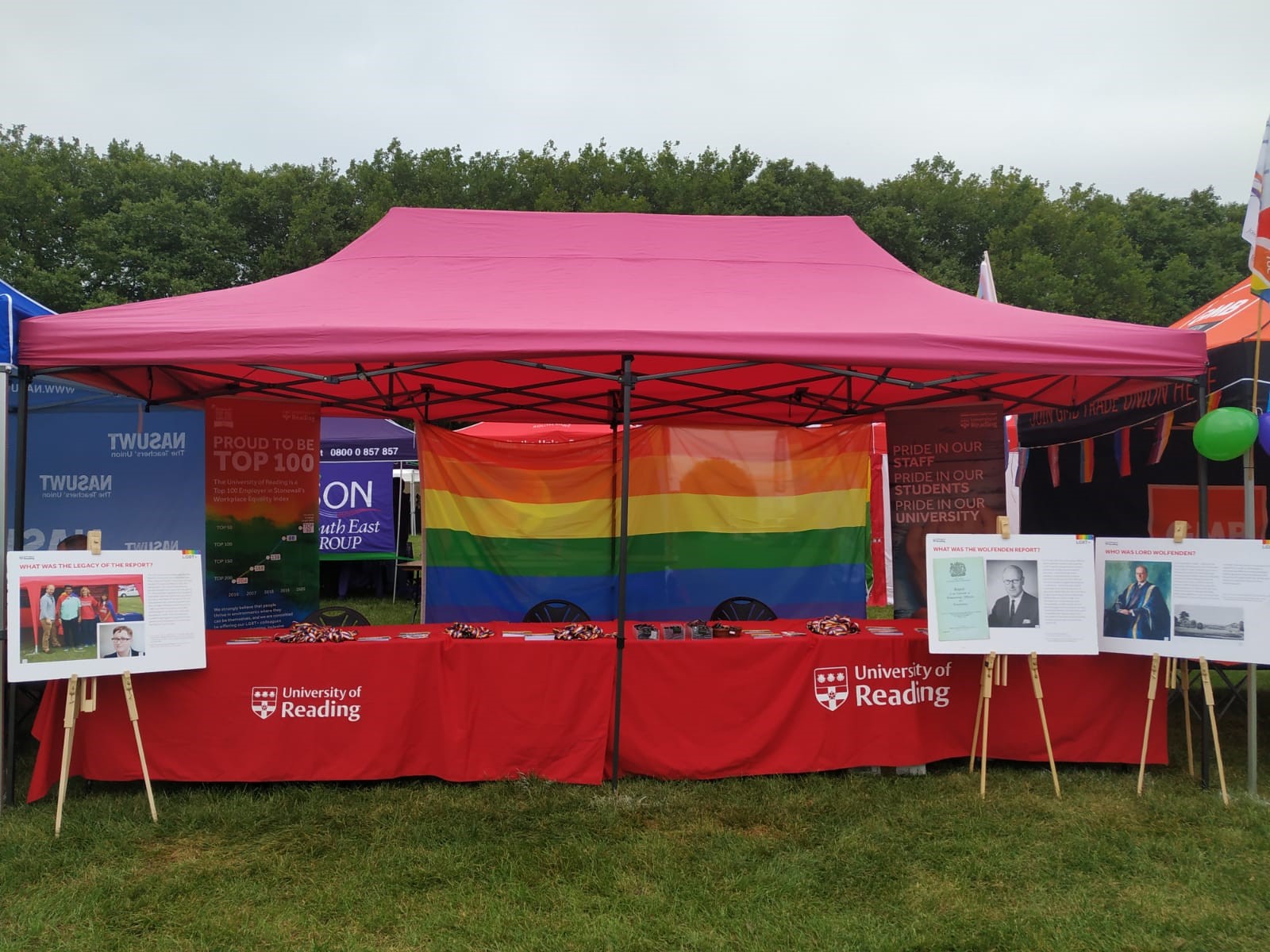 The University's Stand at the Love Unites festival set up, waiting for guests to arrive; A 6 metre by 3 metre Gazebo with a hot pink covering. Two large tables are under the gazebo, covered by the University of Reading tablecloths, in our signature red colour. A large rainbow flag hangs from the back of the Gazebo Three large signs are standing on easels in front of the stand, showing the Lord Wolfenden and the cover of his report. There is text explaining the Wolfenden legacy, and another image in modern day, showing University of Reading. The text describes the modern-day impact of the Wolfenden report on staff, students, and the wider community. 