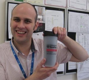 Sam Tyler grins whilst holding travel mug up to the camera