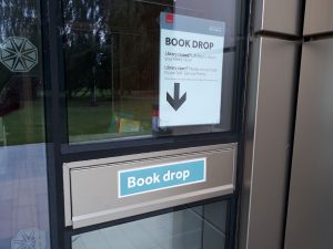 Metal letter-box type flap (labelled Book drop) in glass and metal section of building