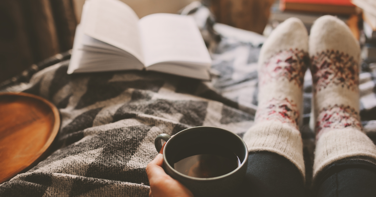 Image of a person sitting on a bed, close-up of feet with cosy socks on, book open on bed. Coffee in hand.