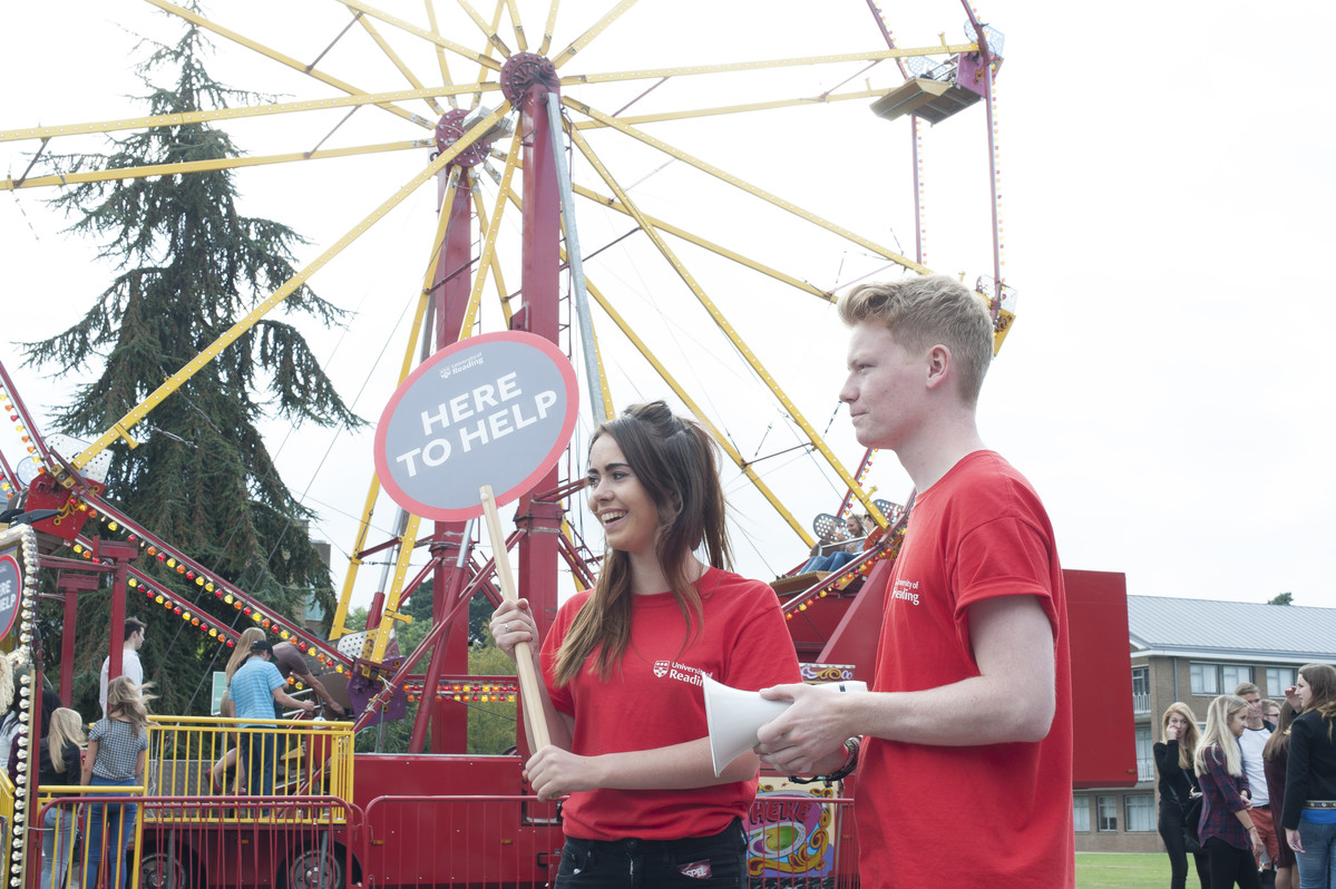 Two student ambassadors with a 'here to help' sign at the Welcome fayre. Standing in front of a fairground wheel.